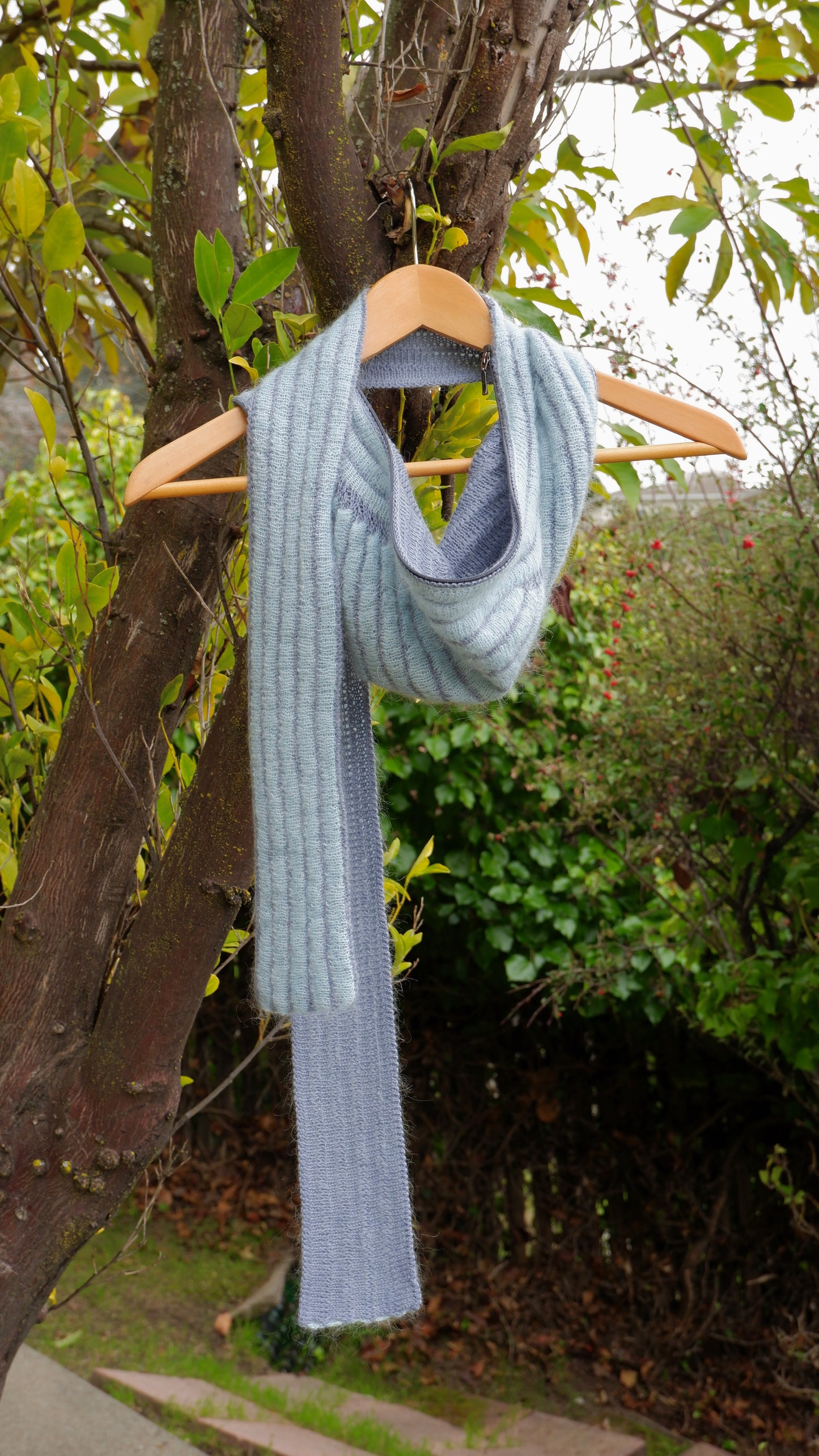 Light blue scarf on a wooden hanger against a natural background with trees and greenery.